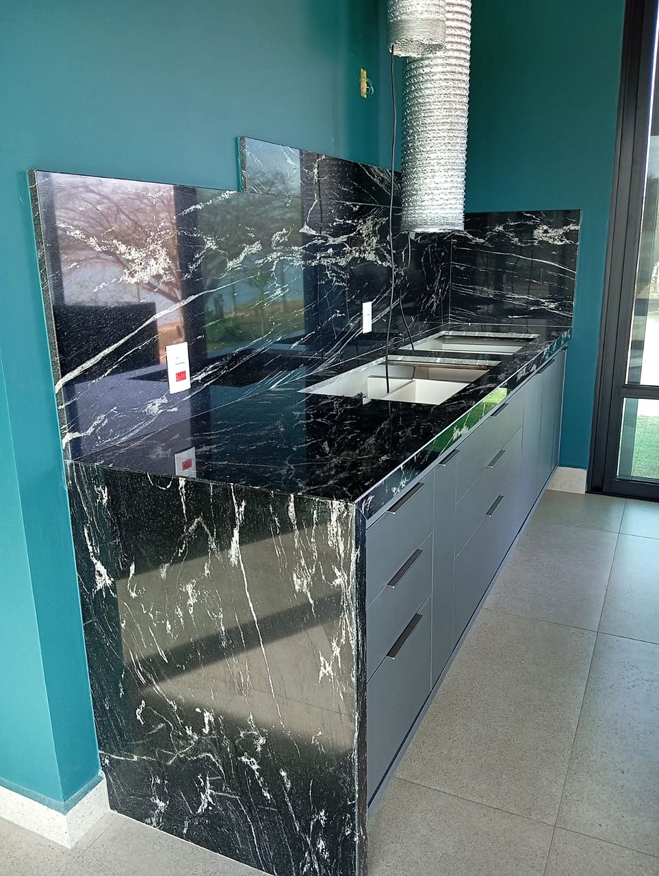 Bancada em granito Preto Via Láctea com frontão alto e cascata lateral, diante de parede verde-azulada